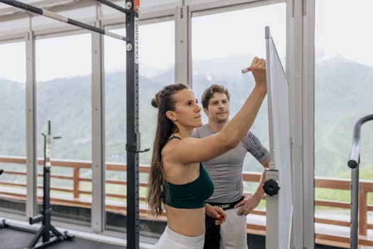 Female fitness coach writes on whiteboard as a male athlete watches in a gym.