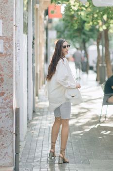 A woman in sunglasses and stylish attire walks down a sunny sidewalk, capturing a blend of fashion and leisure.