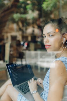Young woman using laptop at a cafe, working remotely with earphones, through a window view.