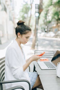 一位年轻女子在户外咖啡馆使用智能手机，桌上放着笔记本电脑和钱包
