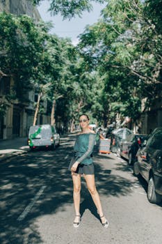 Stylish woman in sunglasses poses confidently on a sunny city street lined with trees.