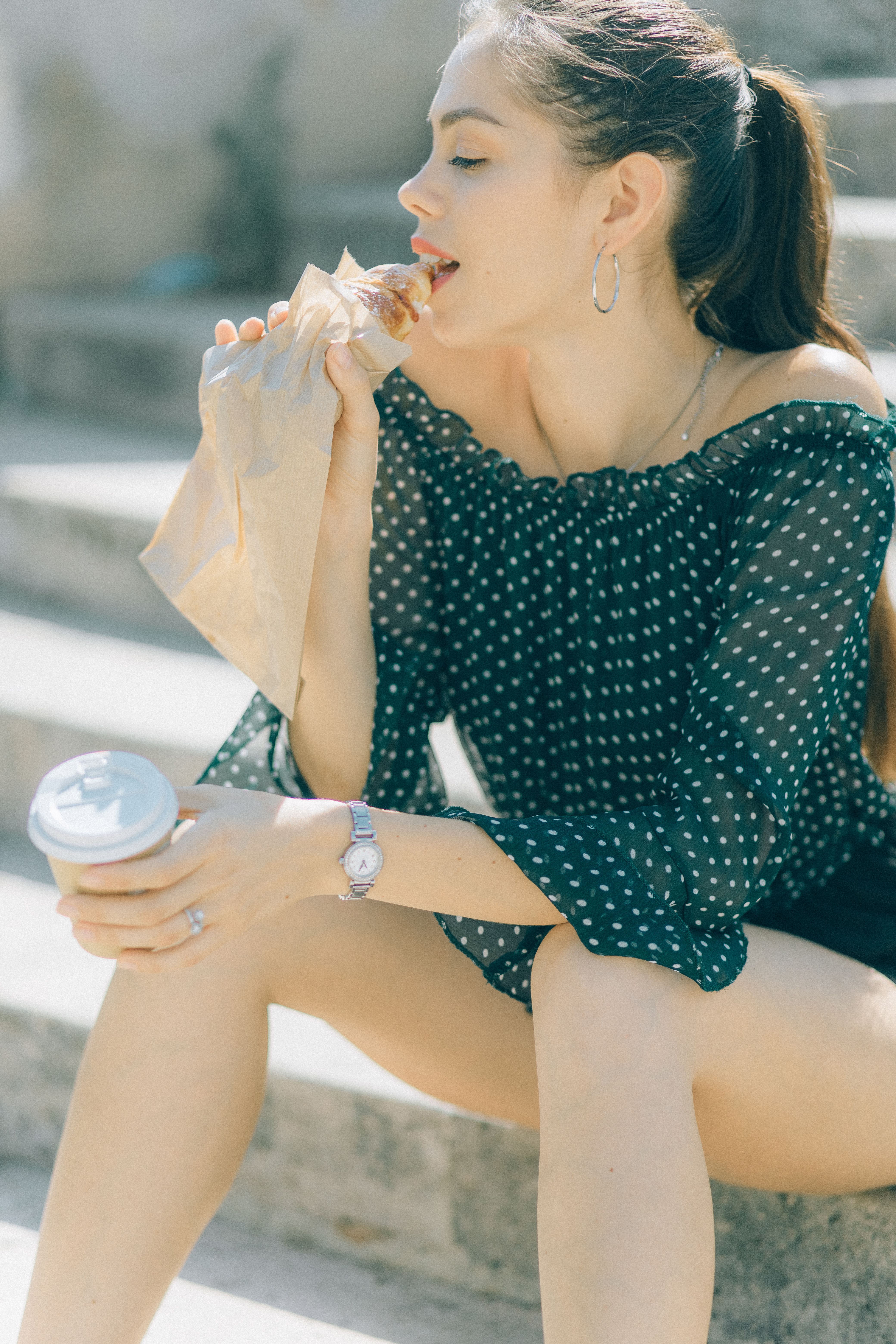 一位穿着圆点衬衫的年轻女子坐在楼梯上，吃着零食，喝着咖啡