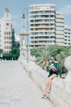 一位别致的女子在阳光明媚的城市景观中，于户外喝着咖啡，将现代时尚与城市建筑融为一体