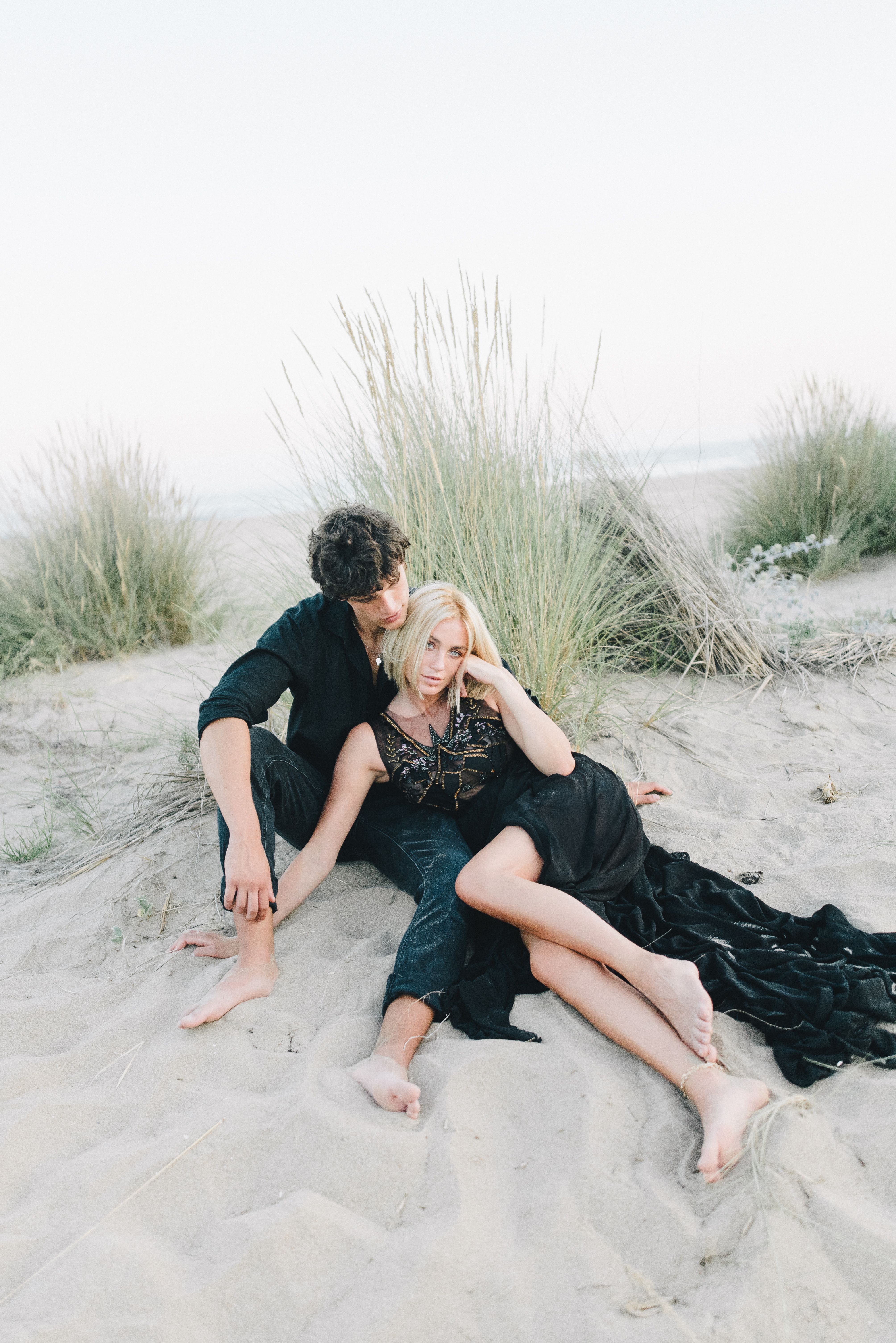 Woman in a Long Black Dress and a Man in Black Long Sleeves Sitting on Sand   Free Stock Photo