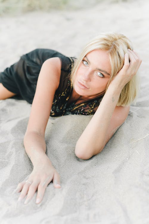 Blonde Woman in Black Dress Lying on Sand on Beach