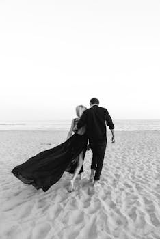 Black and white photo of a couple walking on the beach, capturing a serene romantic moment.