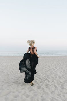 A woman with blond hair in a flowing black gown runs joyfully on a serene beach.