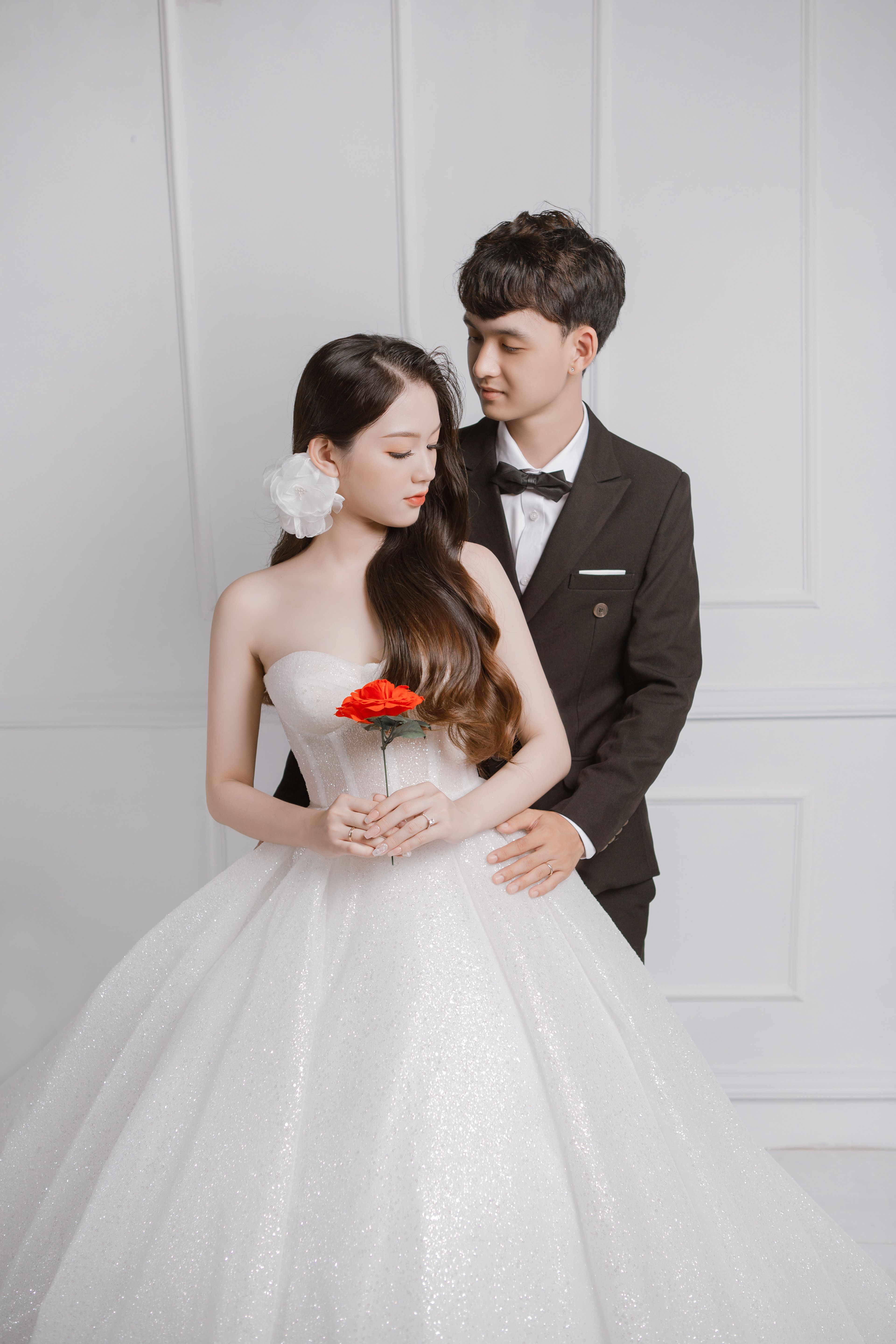 A bride and groom in elegant attire embracing indoors with a red flower.