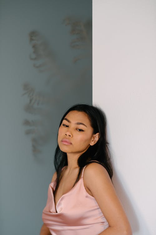 Free Contemplative young Asian female in stylish silk dress standing against white wall in light room and looking away in deep thoughts Stock Photo