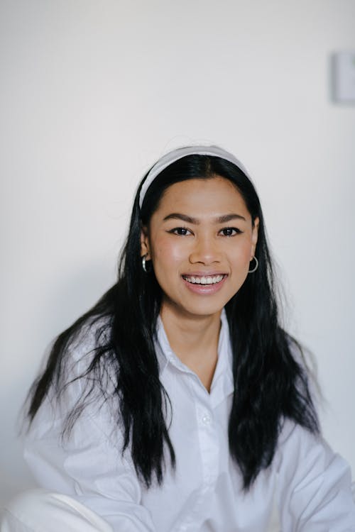 Cheerful young Asian female in white blouse and headband sitting in light room and looking at camera with charming smile