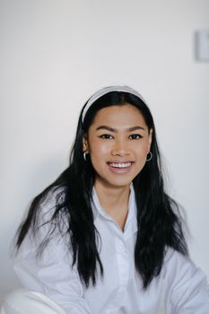 一位快乐的年轻亚洲女性，穿着白色衬衫、戴着头带，坐在明亮的房间里，带着迷人的微笑看向镜头