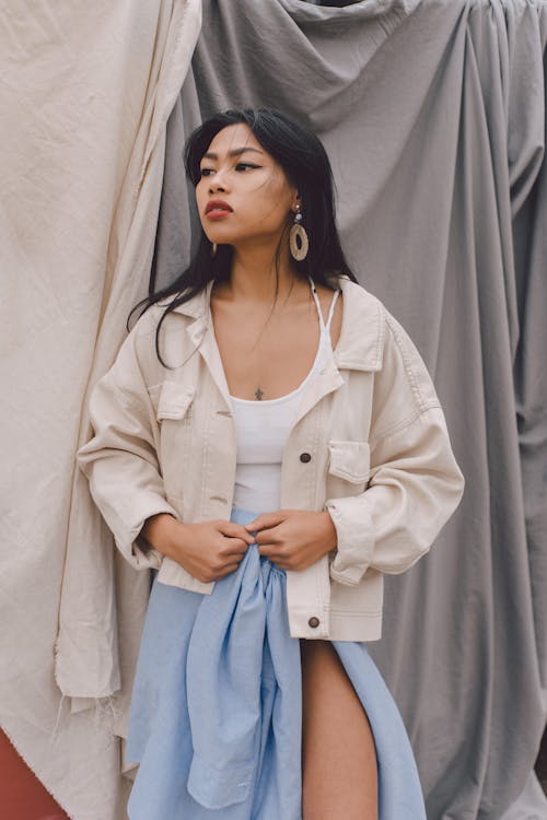 Beautiful young Asian female wearing trendy summer outfit and denim jacket standing against hanging bedsheets and looking away dreamily