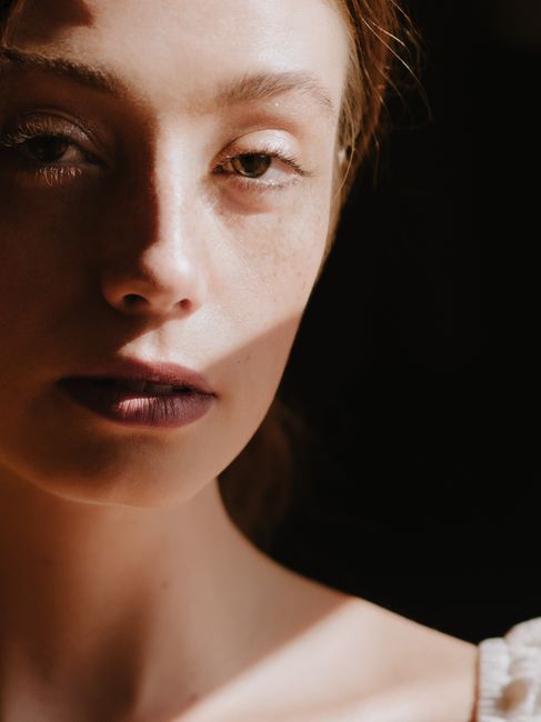 Crop serene pretty female standing in dark room in sunlight and looking at camera with languid glance