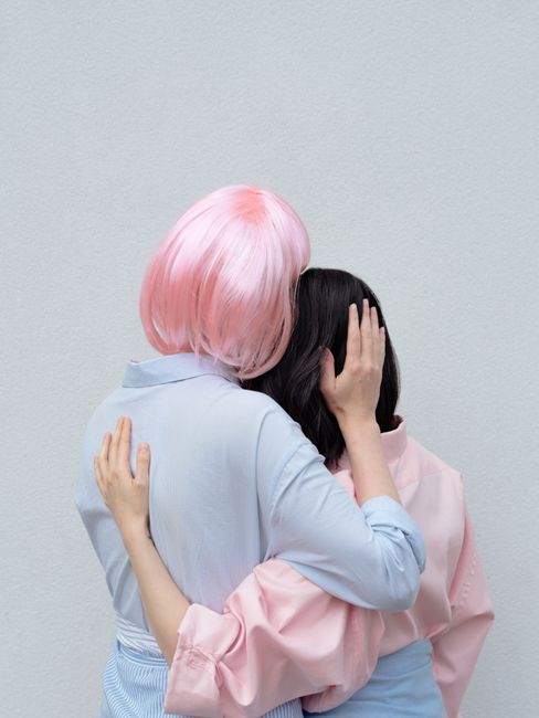 Back view unrecognizable female in blue wear and pink wig hugging girlfriend against gray wall in light studio