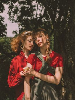 一位平静的女人手握苹果，站在树枝阴影下的树旁，位于她平静的女友身后