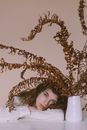 Young tender female looking at camera and resting under wilted plant with leaves in vase