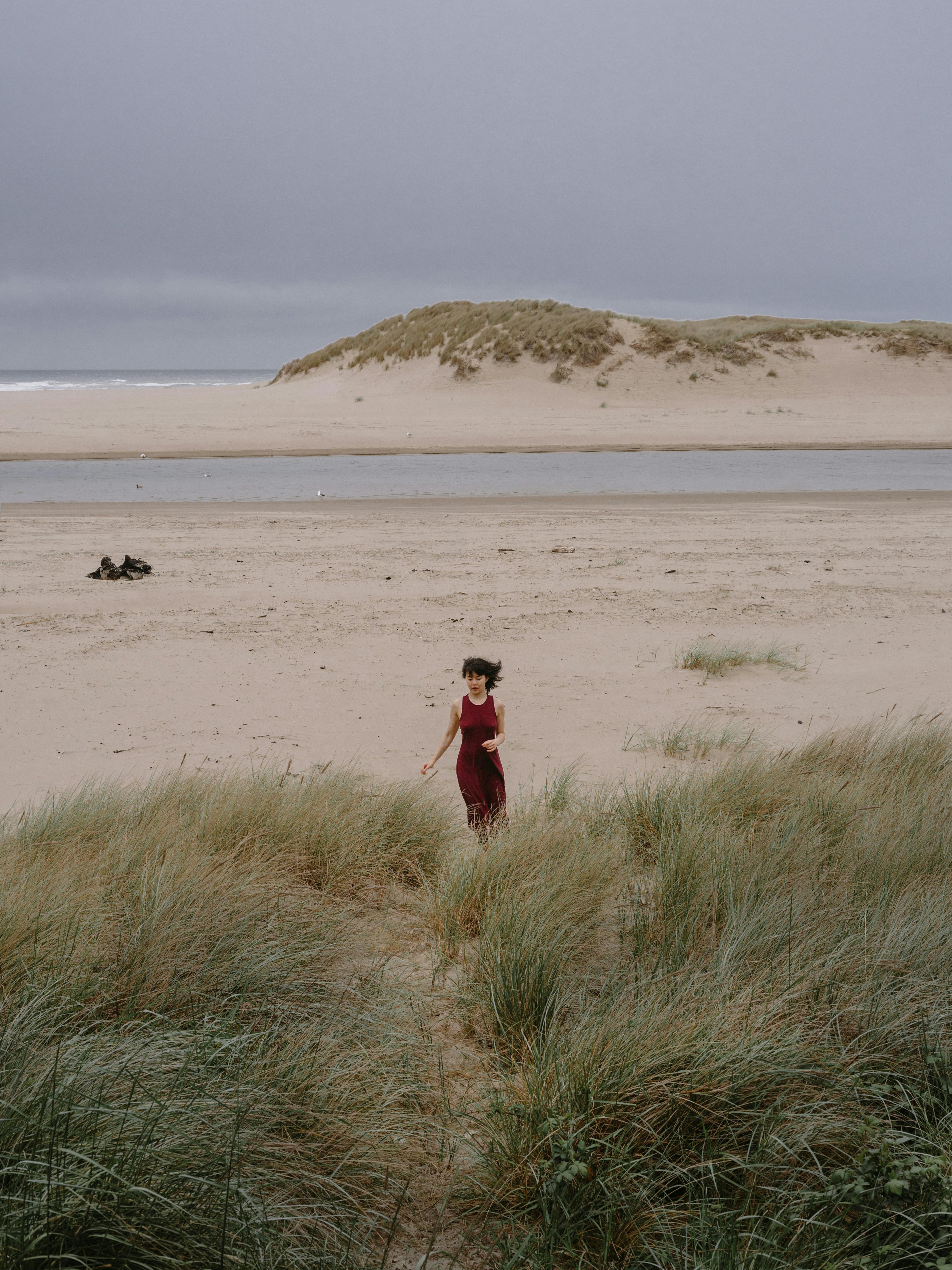 Young slim anonymous female in red dress promenading on sandy coast of ocean with grass in windy cloudy weather