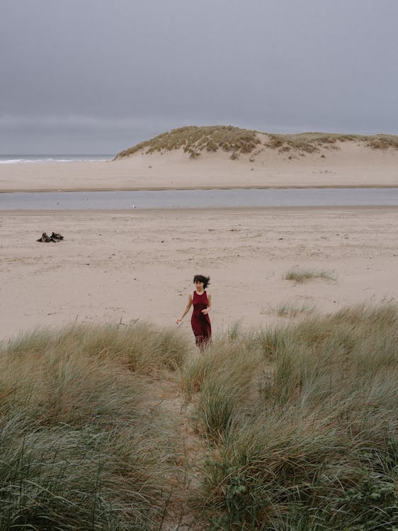 Free Young slim anonymous female in red dress promenading on sandy coast of ocean with grass in windy cloudy weather Stock Photo