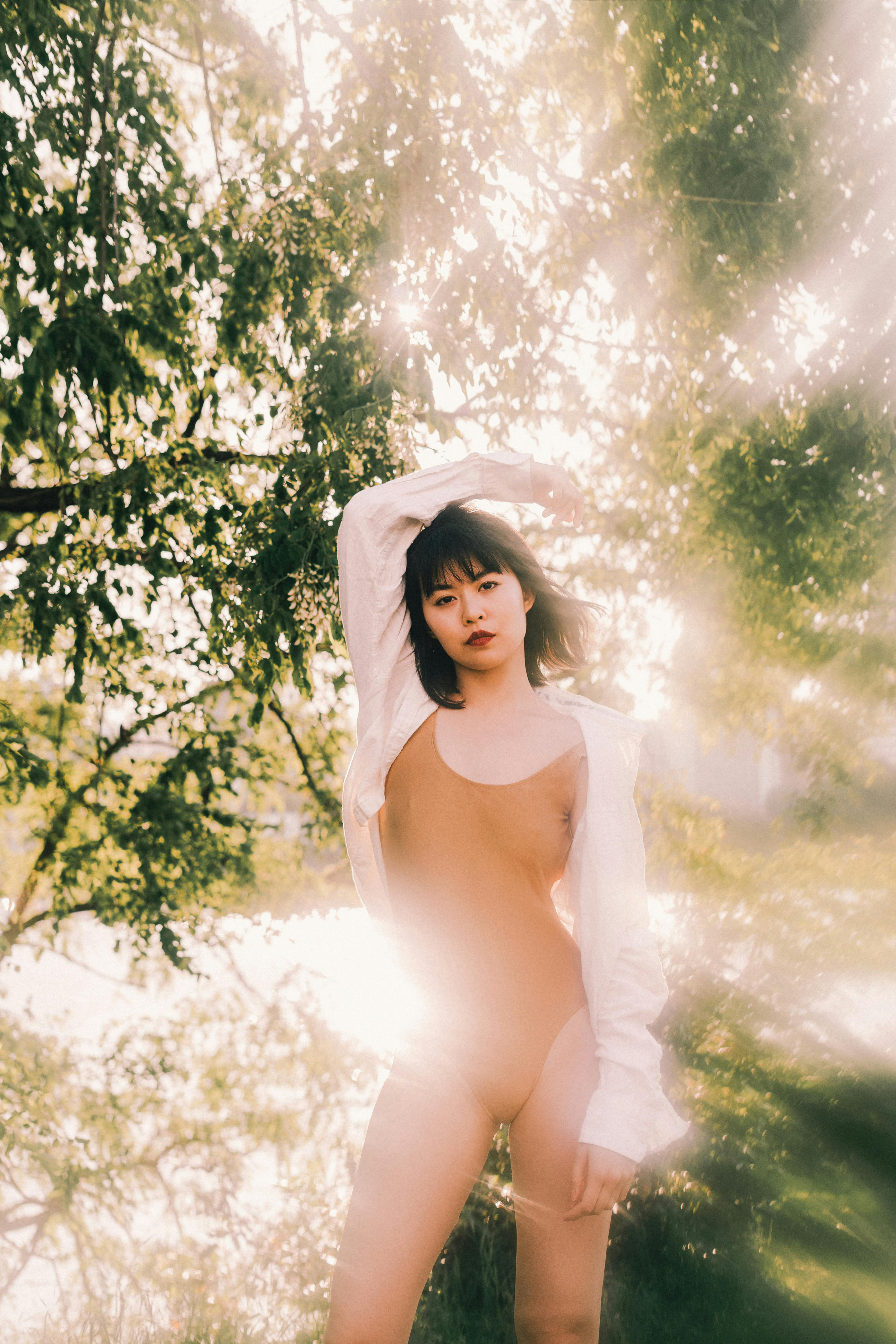 Young content ethnic female with hand on head looking at camera in green forest in back lit