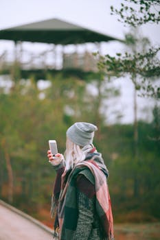 A young woman in cozy attire using her smartphone in a tranquil forest setting, perfect for nature-themed lifestyle imagery.