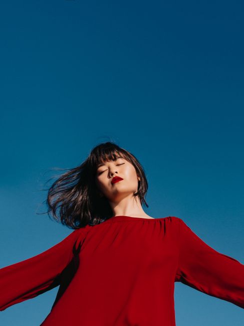 From below of young Asian female in red blouse with hands sideways and eyes closed against cloudless blue sky