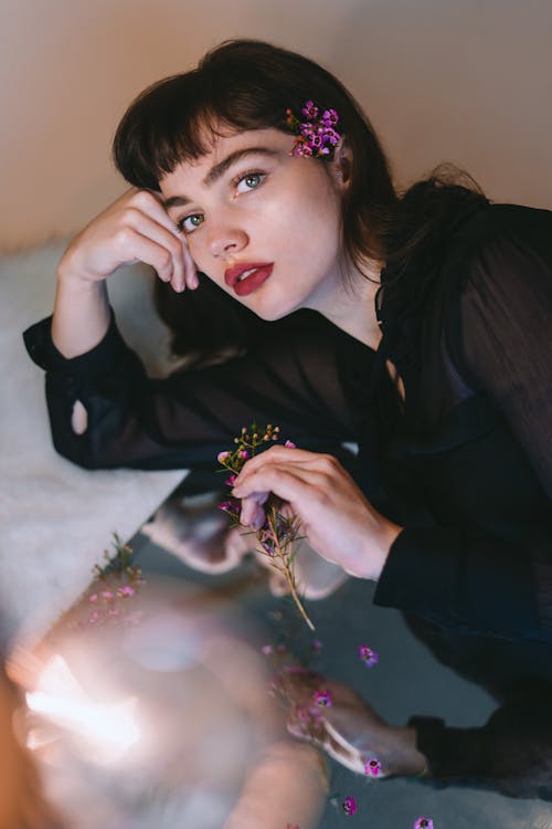 Free High angle of young dreamy female in black clothes with flower in hand and behind ear leaning on mirror surface and looking at camera Stock Photo