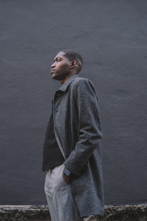 Side view of serious young African American male in formal clothes with hands in pockets standing near dark gray wall