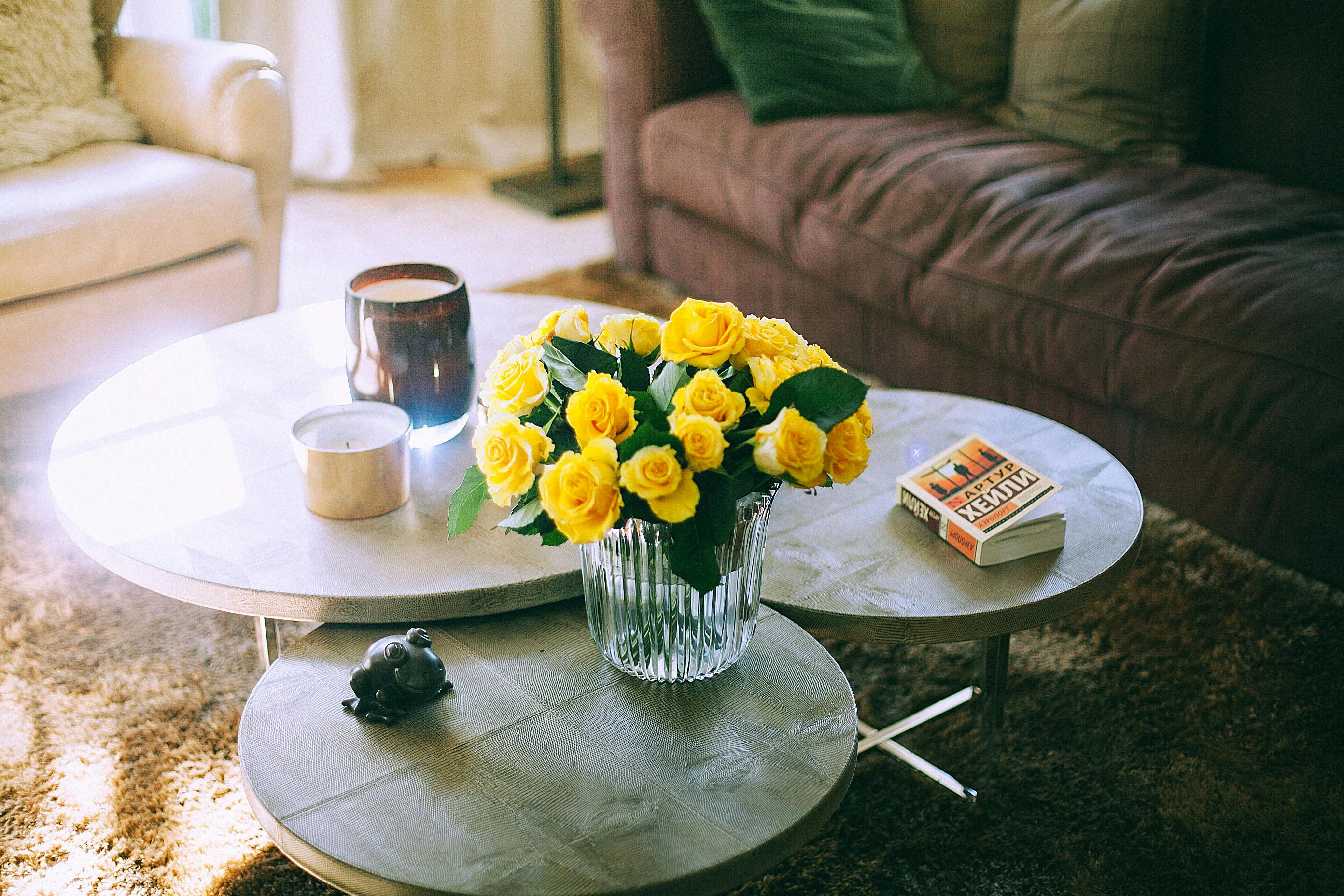 Modern table with vase of yellow roses and candles with book near couch and armchair on carpet in room