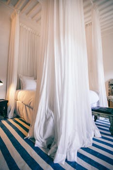 Spacious bedroom with large bed with white curtains hanging on ceiling in daylight