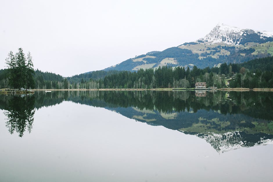 Picturesque landscape of calm lake reflecting rural house on bank and forest in front of mountain ridge covered with snow on cloudy day