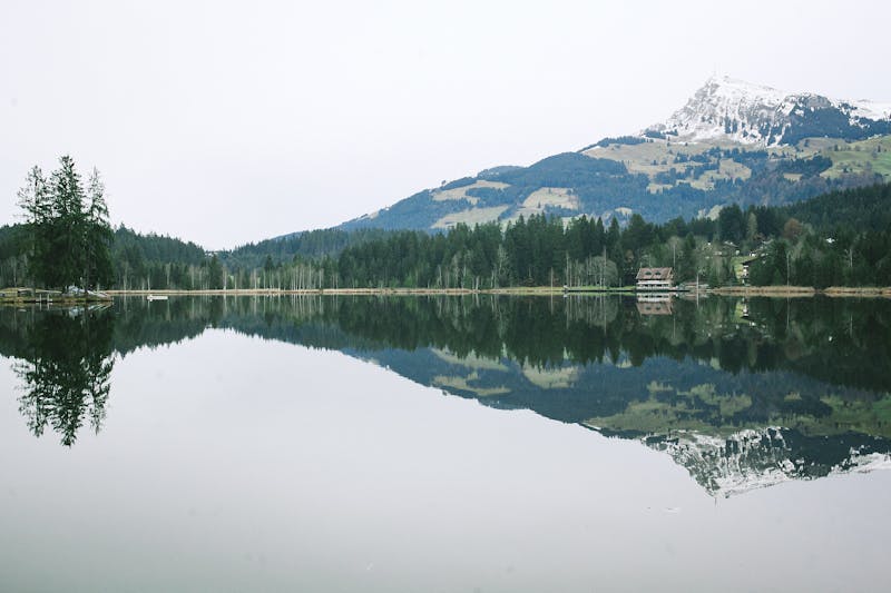 Picturesque landscape of calm lake reflecting rural house on bank and forest in front of mountain ridge covered with snow on cloudy day