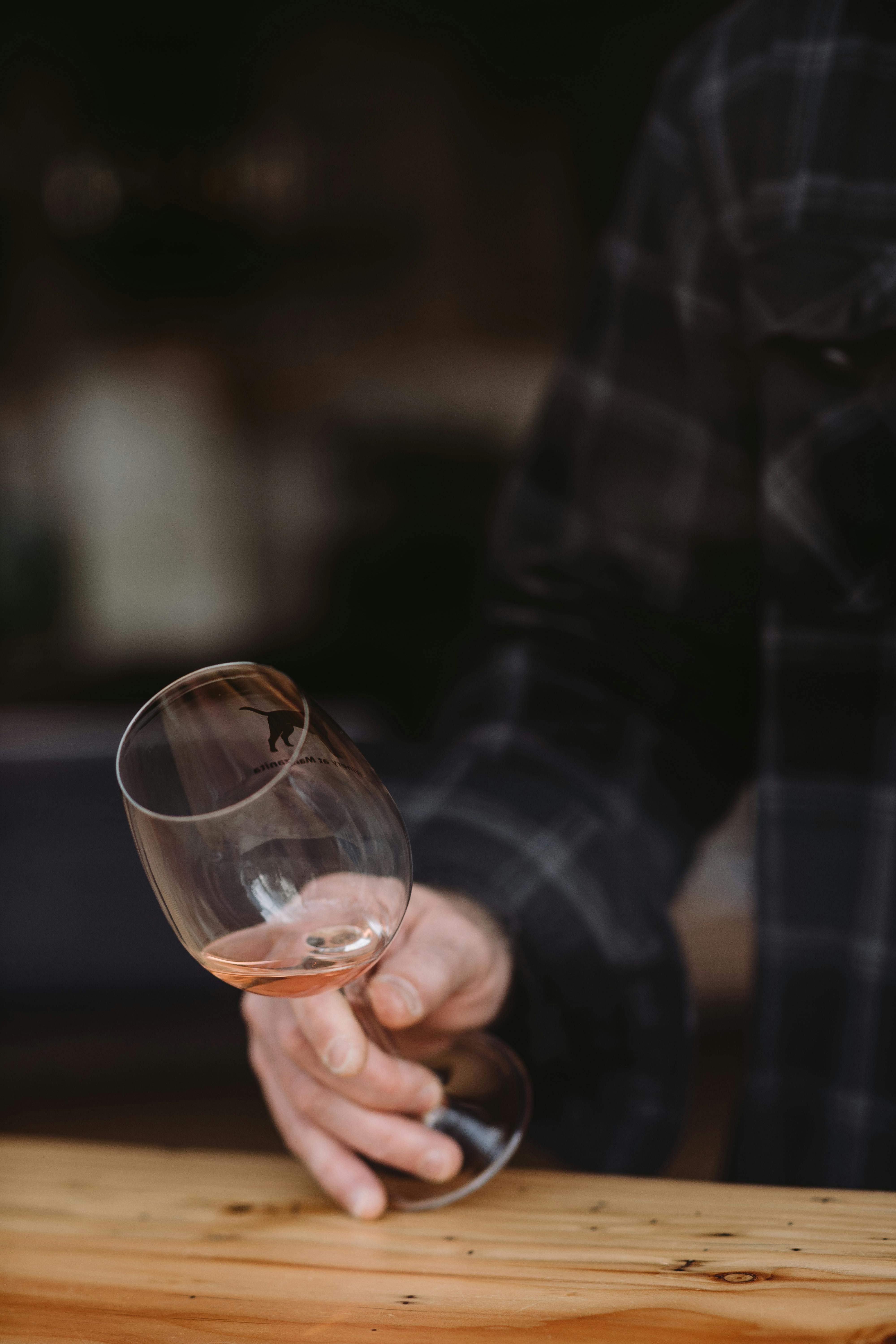 Unrecognizable man in casual shirt holding glass with white wine sitting at wooden table on blurred background