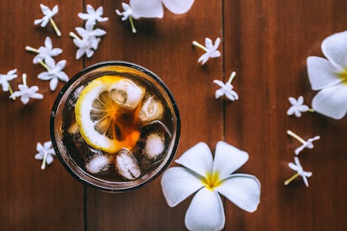 Top view of glass with fresh drink with slice of lemon and ice cubes on wooden background with plumeria flowers