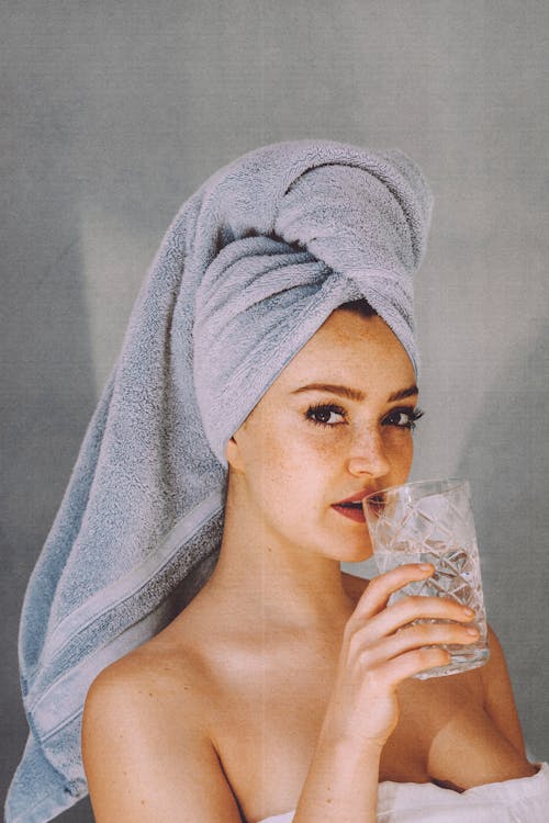 Young woman with hair wrapped with towel and bared shoulders drinking water and looking at camera
