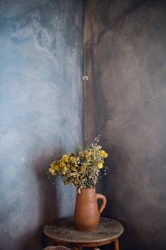 A rustic display of dried yellow flowers in a terracotta vase on a wooden table against a textured wall.