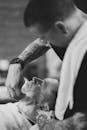 Black and white side view of tattooed barber wearing eyeglasses applying shaving foam on customer face before shaving in barbershop