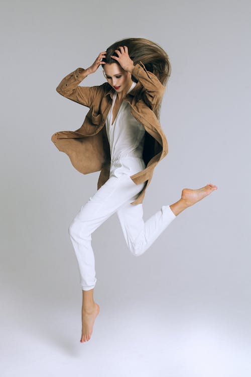 Full body of young stylish barefoot female in trendy clothes touching head while jumping on white background