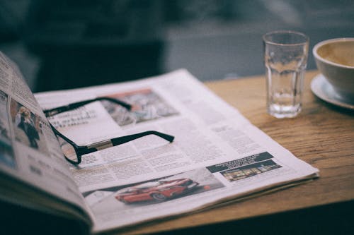 Eyewear on magazine with text in columns and illustrations near cup with saucer on wooden table in cafeteria