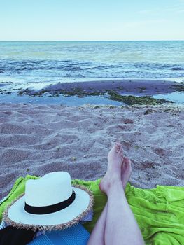 Crop barefoot person with straw hat lying on green plaid on sandy shore near blue waving sea