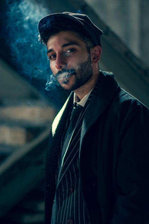 Free A stylish man in London wearing a black coat and hat, exhaling smoke in a moody, urban setting. Stock Photo