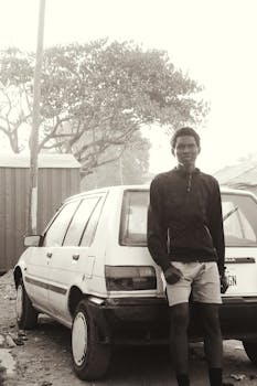 一位身材修长的非裔美国人，身着黑白装束，神情冷漠，白天在院子里倚靠着一辆老式简约的白色汽车，望向镜头