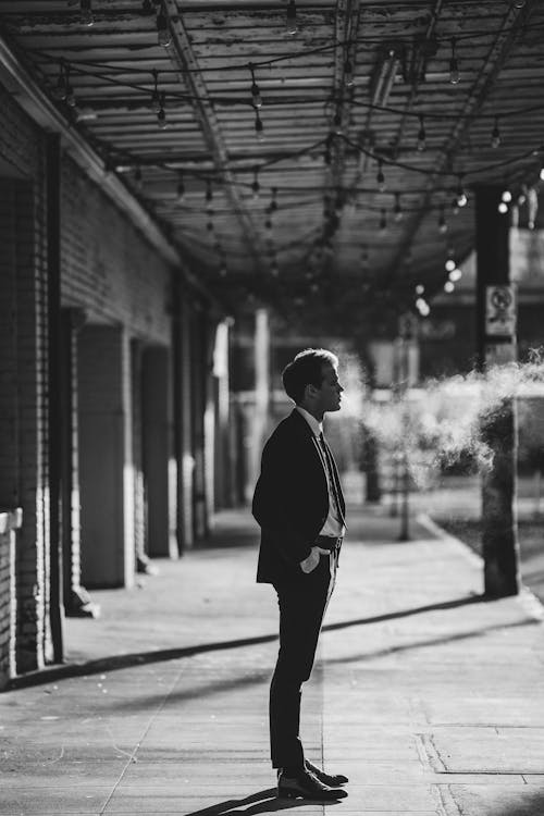 Free Black and white side view of male entrepreneur in formal suit with hand in pocket looking forward between haze and building on pathway Stock Photo