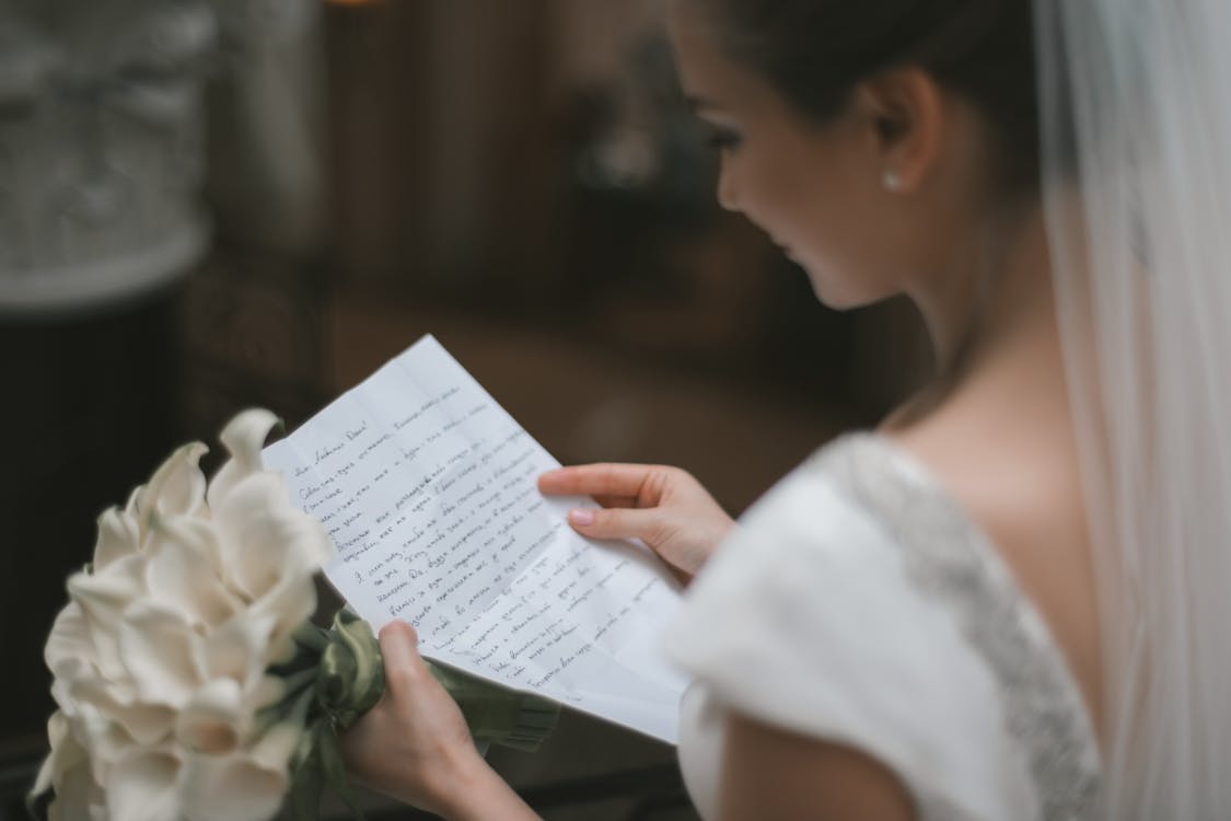 Free Back view of crop charming bride in white dress and veil reading wedding vow on paper sheet with bouquet of arum lilies during weeding ceremony Stock Photo