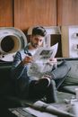 Handsome bearded man in jean outfit focusing on reading newspaper while sitting on comfortable couch in common room with big decorative letters on wall