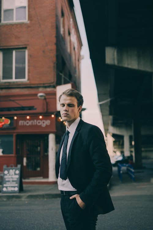 Confident young man in formal suit standing on city street with closed eyes and keeping hands in pocket