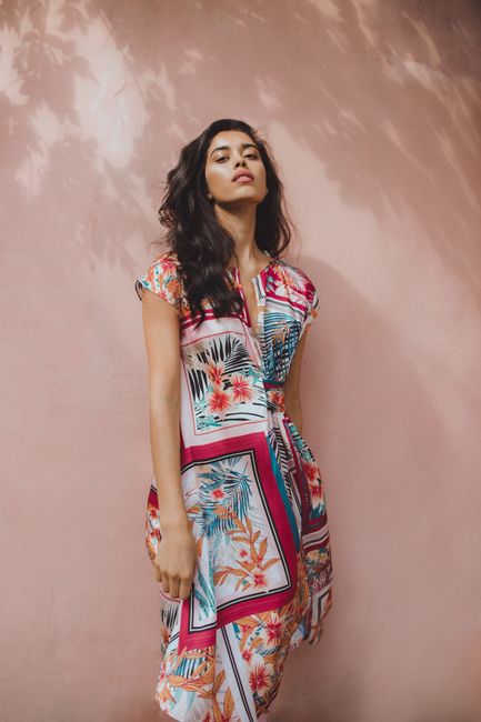 Low angle of young Hispanic female in summer colourful printed dress standing beside wall and looking at camera