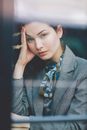 Through glass of young contemplative female in elegant suit leaning on hand and looking at camera while sitting at table in cafe