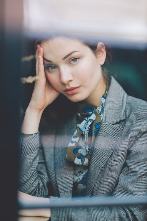 Free Through glass of young contemplative female in elegant suit leaning on hand and looking at camera while sitting at table in cafe Stock Photo