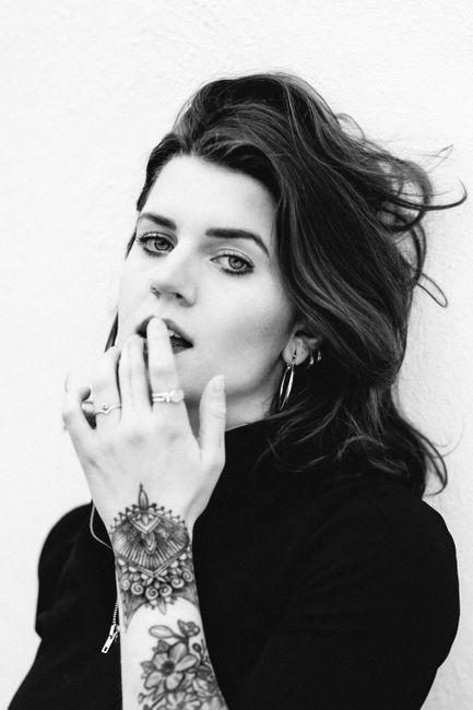 Young serious female in dark clothes looking at camera touching lips with tattooed hand