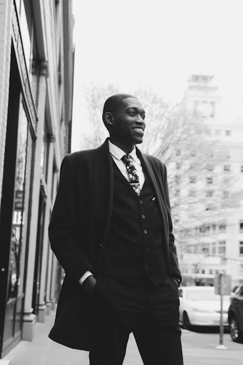 Free Low angle of young cheerful African American man in elegant suit and coat standing on city street keeping hand in pocket and looking away Stock Photo